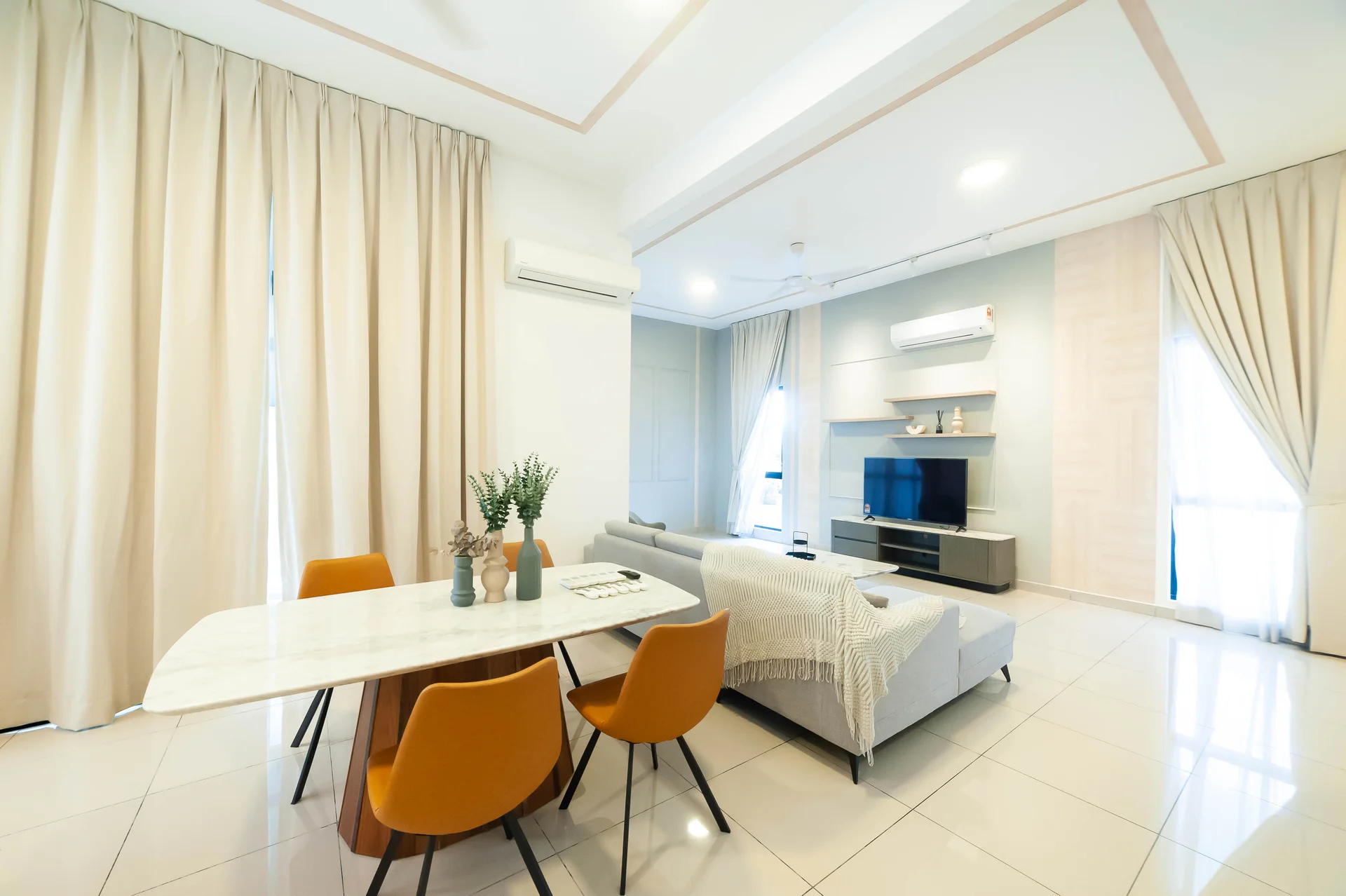 Living features: marble dining table, 4 orange upholstered dining chairs, light grey fabric sofa, TV console, wall-mounted TV, recessed downlights, floor-to-ceiling curtains, cream painted walls, white ceiling with crown moulding, air conditioning unit, decorative shelves, decorative vases; style: modern.