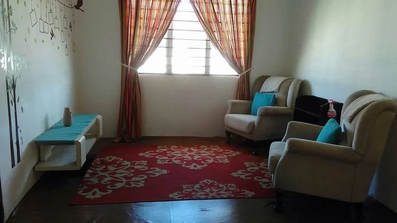 Living features: two beige armchairs, blue accent cushions, red area rug with white pattern, light wood console table, window with horizontal bars, curtains with orange and yellow stripes, decorative wall decals, wooden flooring.