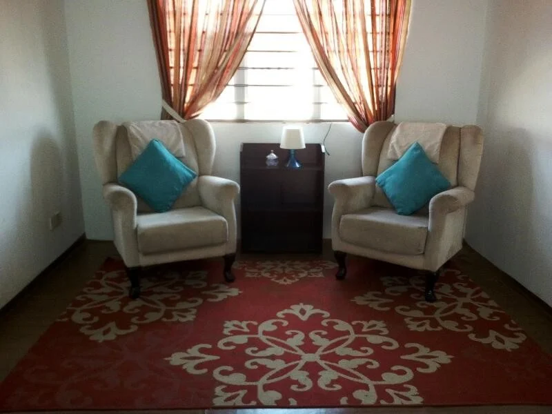 Living features: two beige armchairs with wingback design, blue decorative cushions on chairs, small wooden side table, table lamp on side table, red area rug with white floral pattern, curtains with orange and cream stripes, wooden flooring, window with horizontal bars; style: contemporary.