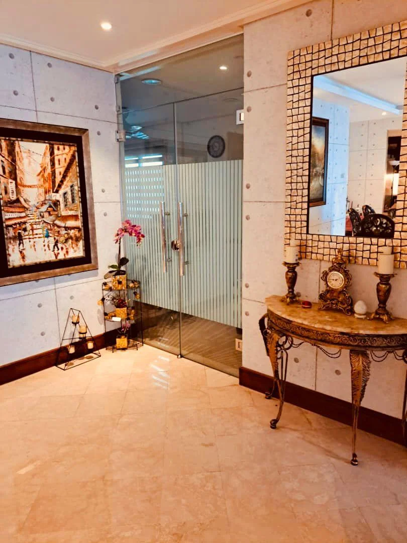 Room features: grey concrete walls, marble tile flooring, glass door, decorative mirror with mosaic frame, framed artwork, console table with ornate legs, table clock, decorative vase with flowers, wall-mounted clock, ambient lighting; style: modern.