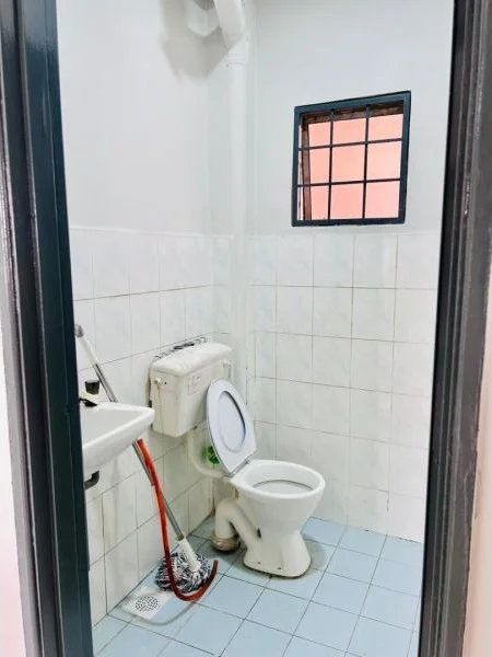Bathroom features: white tiled walls, light blue tiled flooring, one-piece toilet, wall-mounted sink, window with black metal grille, exposed plumbing, toilet paper holder, cleaning supplies visible.
