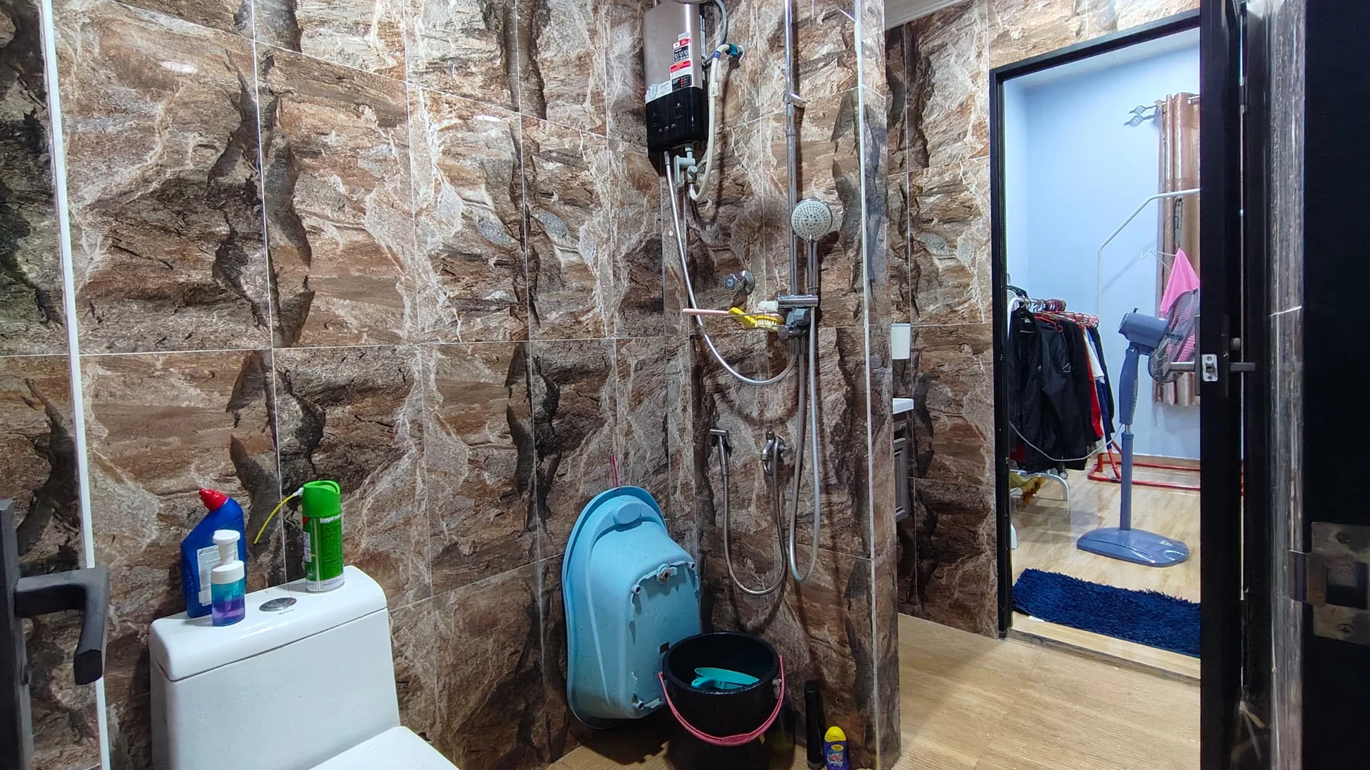 Bathroom features: one-piece toilet, shower area with glass partition, rain shower head, handheld shower, tile cladding with stone pattern, wooden flooring, shower caddy, bucket, cleaning supplies, wall-mounted water heater; style: modern. Room features: clothes rack, wooden flooring, blue area rug.
