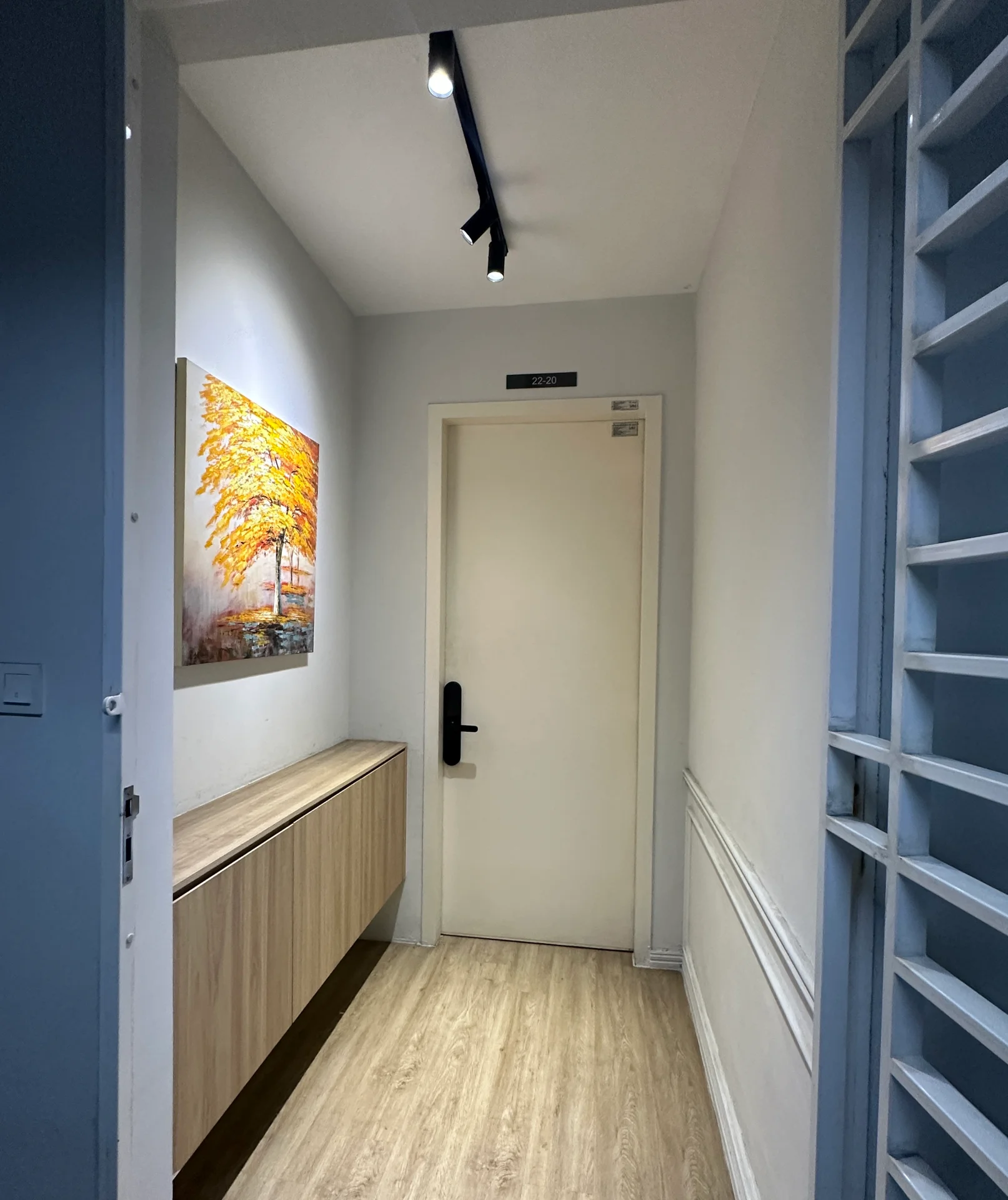 Room features: light wood flooring, white painted walls, wall-mounted artwork with autumn tree, wooden console table, track lighting, door with black handle, decorative wall paneling; style: modern.