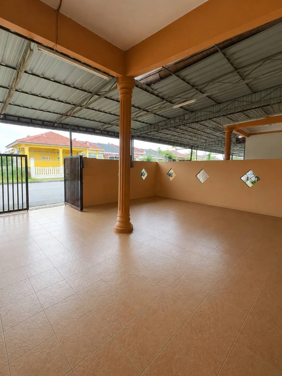 Exterior features: covered porch area, tan tiled flooring, orange painted columns, metal roof, gated entrance, decorative diamond-shaped windows, plain wall with orange paint, open space, surrounded by greenery; style: modern.