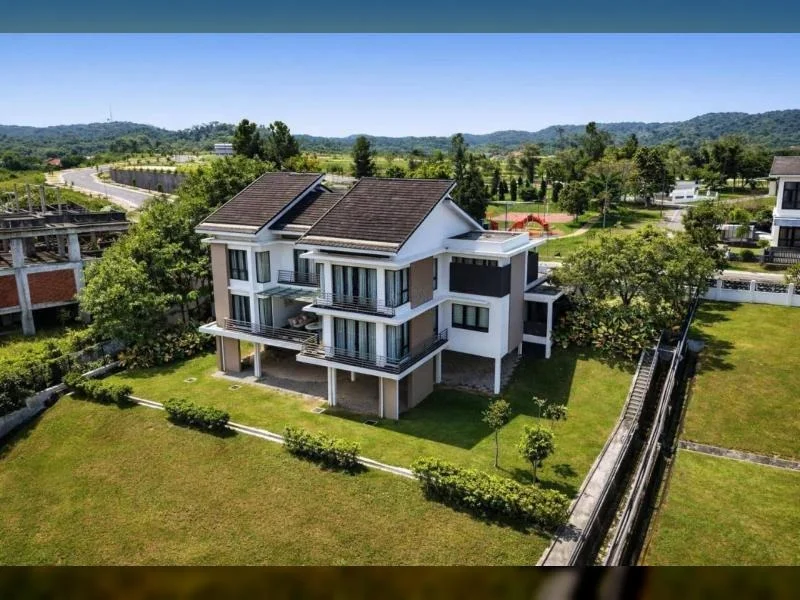 Exterior type: detached bungalow; features: double-storey, white painted facade, dark brown pitched tile roof, projecting balconies, glass balcony railings, large windows, covered ground-floor terrace, surrounded by mature greenery, manicured lawn, stone-paved walkway, adjacent open green field, low-rise residential setting; style: contemporary.