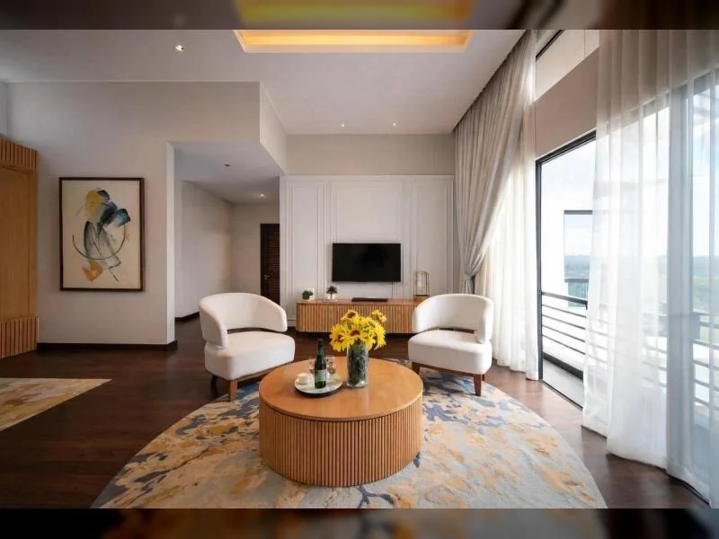 Living features: white painted walls, dark wood flooring, recessed ceiling downlights, tray ceiling with warm cove lighting, floor-to-ceiling windows, sheer curtains, floor-length drapes, sliding glass door to balcony, balcony railing, wall-mounted TV, wood TV console, 2 white upholstered accent chairs, round wooden coffee table, large area rug with abstract pattern, framed abstract artwork, built-in wood slat feature panel, minimalist decor; style: modern; view: garden.