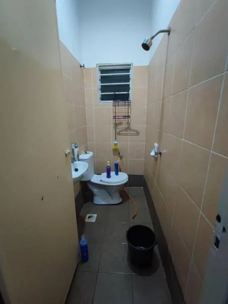 Bathroom features: ceramic tile flooring, beige tiled walls, white toilet, shower head, wall-mounted shower, window with metal grille, plumbing fixtures visible, cleaning supplies, bucket.