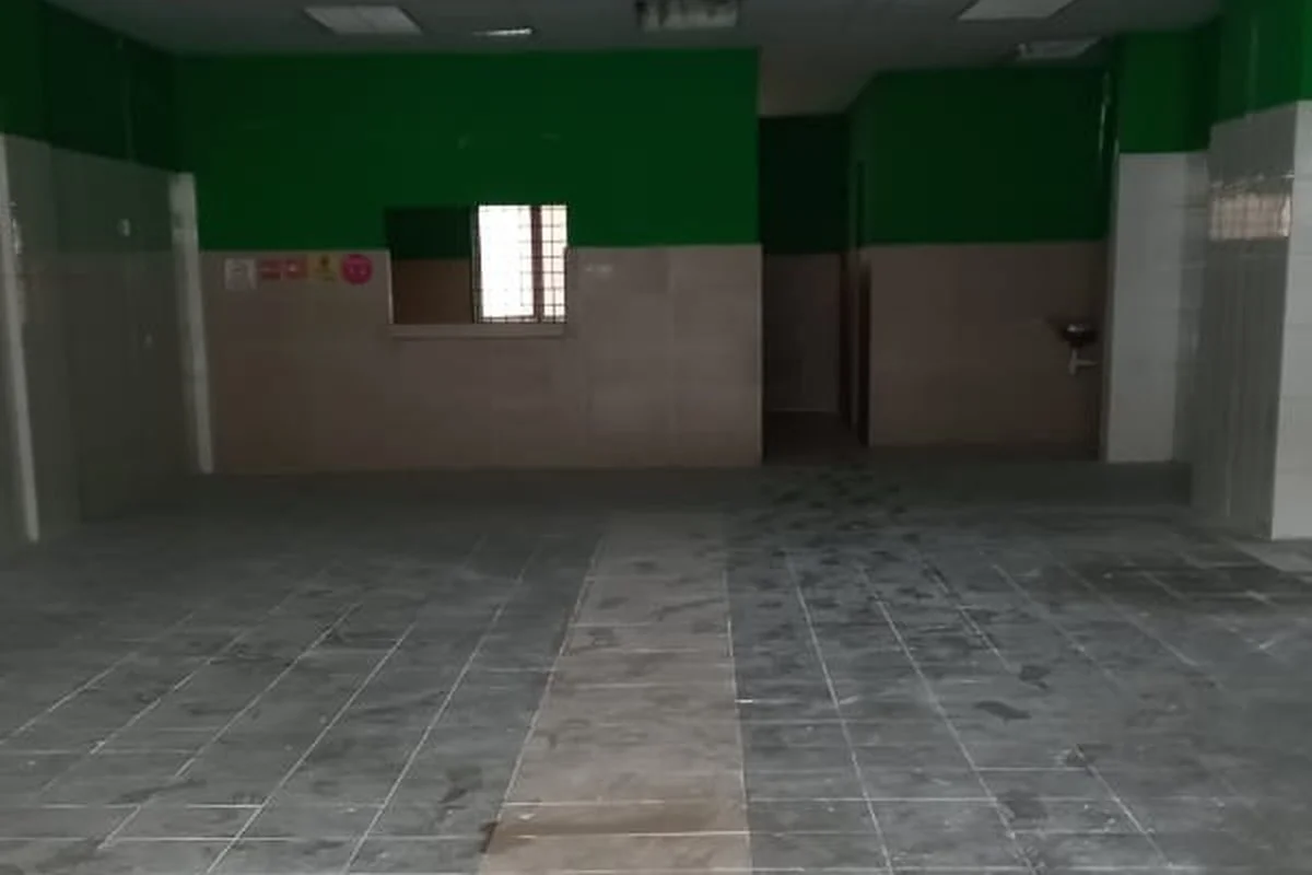 Room features: green painted walls, light beige tiled walls, grey tile flooring, large open space, window with grille, sink on wall, fluorescent lighting, no visible furniture or fixtures.