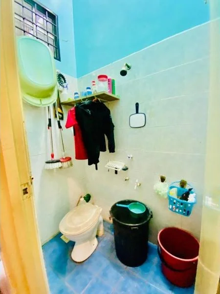 Bathroom features: white ceramic toilet, blue tiled flooring, light blue painted walls, shelf with cleaning supplies, mirror, black trash bin, red bucket, hanging clothes, shower head, wall-mounted faucet.