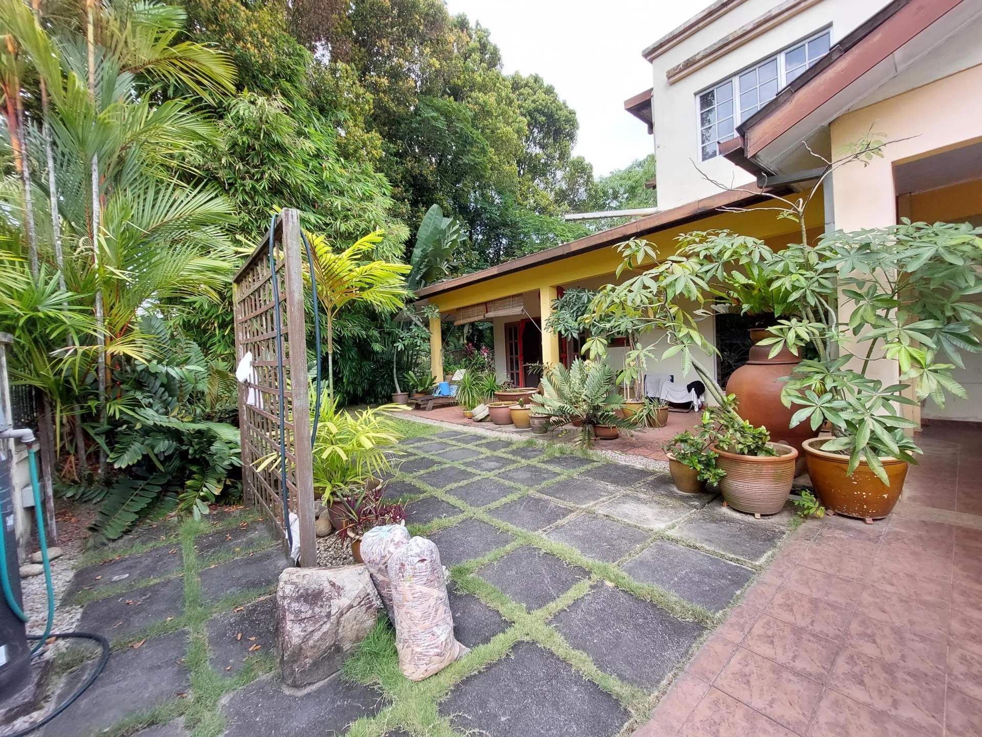 Exterior type: detached bungalow; features: manicured lawn, tropical landscaping, paved walkway, various potted plants, large clay pots, stone pathway, covered porch, green foliage, garden area; style: tropical-modern.