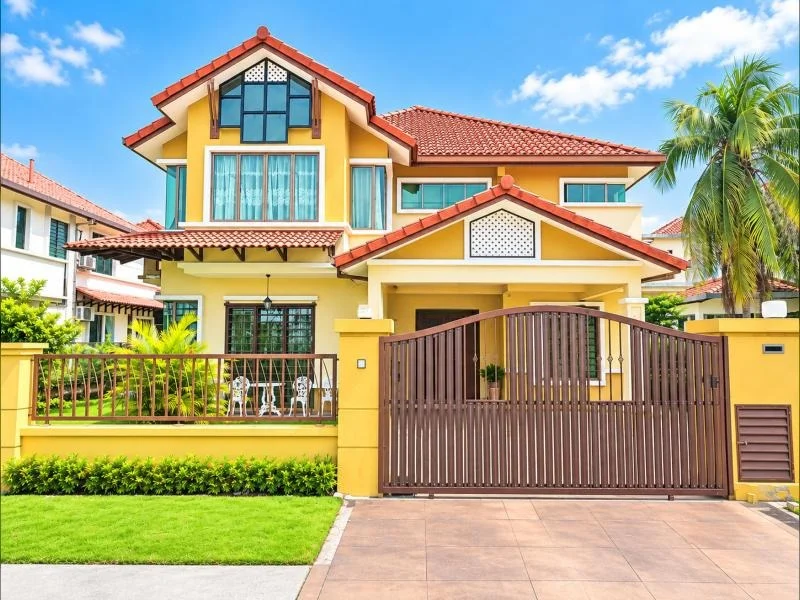 Exterior type: detached bungalow; features: double-storey, yellow painted facade, white trim facade, red tiled pitched roof, projecting gables, decorative perforated gable panels, large front balcony, covered porch, brown metal sliding gate, car porch with paved driveway, stone-paved front yard, manicured lawn, tropical landscaping, palm trees, fronts a quiet residential street; style: tropical-modern.