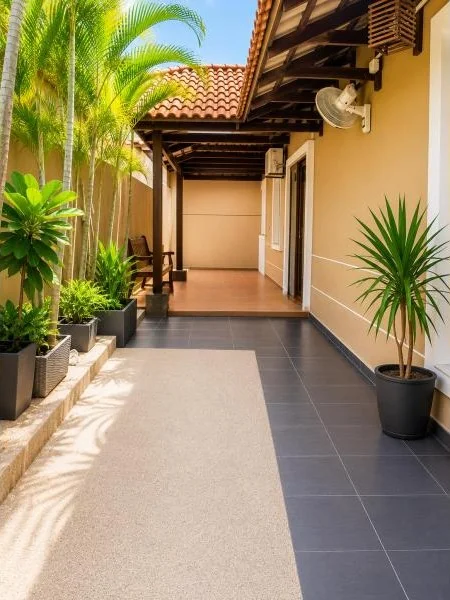 Exterior type: linked terrace house; features: single-storey, pitched tile roof, terracotta roof tiles, exposed timber roof structure, cream painted facade, covered side veranda, dark grey floor tiles, light beige outdoor flooring, sliding glass door, white-framed windows, wall-mounted oscillating fan, air-conditioning unit, potted tropical plants, stone planter border, stone-paved walkway, fronts a quiet residential street; style: traditional.