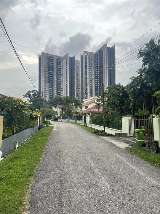 Exterior type: high-rise condominium; features: modern glass facade, multiple balconies, surrounded by greenery, paved road leading to the building, street lighting, manicured landscaping, adjacent residential houses, power lines visible; style: modern.