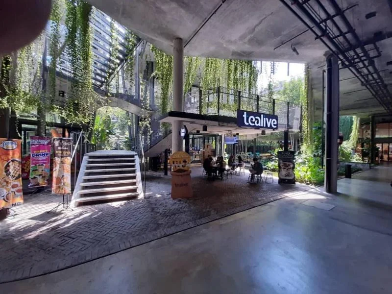 Exterior type: commercial space; features: open-air design, concrete flooring, greenery with hanging plants, staircase leading to upper level, outdoor seating area, signage for tea shop, decorative planters, large windows, exposed ceiling, modern architectural elements; style: modern.