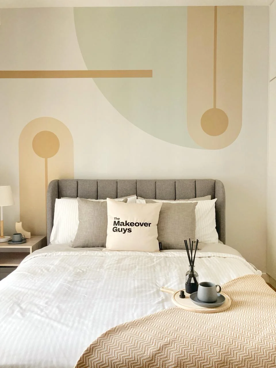 Bedroom features: king-size bed with grey upholstered headboard, white bedding with striped pattern, decorative throw blanket in beige, two grey and beige decorative pillows, nightstand with lamp, framed artwork on wall, wall with abstract geometric design in muted colors, wooden tray with coffee cup and decorative items, reed diffuser, flooring not visible; style: modern.