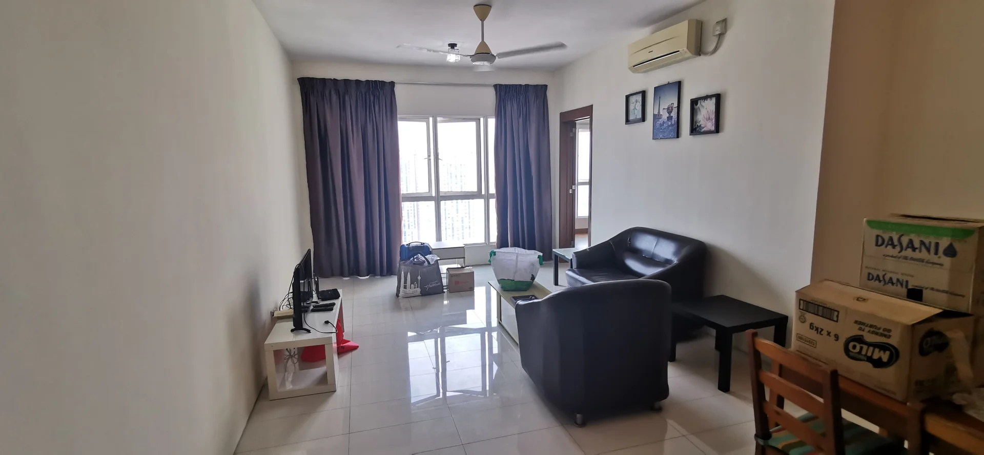 Living features: white painted walls, glossy tile flooring, 5-blade ceiling fan, wall-mounted air conditioning unit, floor-to-ceiling windows, black leather sofa, coffee table, TV console, wall-mounted TV, decorative framed artwork, curtains, cardboard boxes, wooden dining chairs; style: modern; view: city.