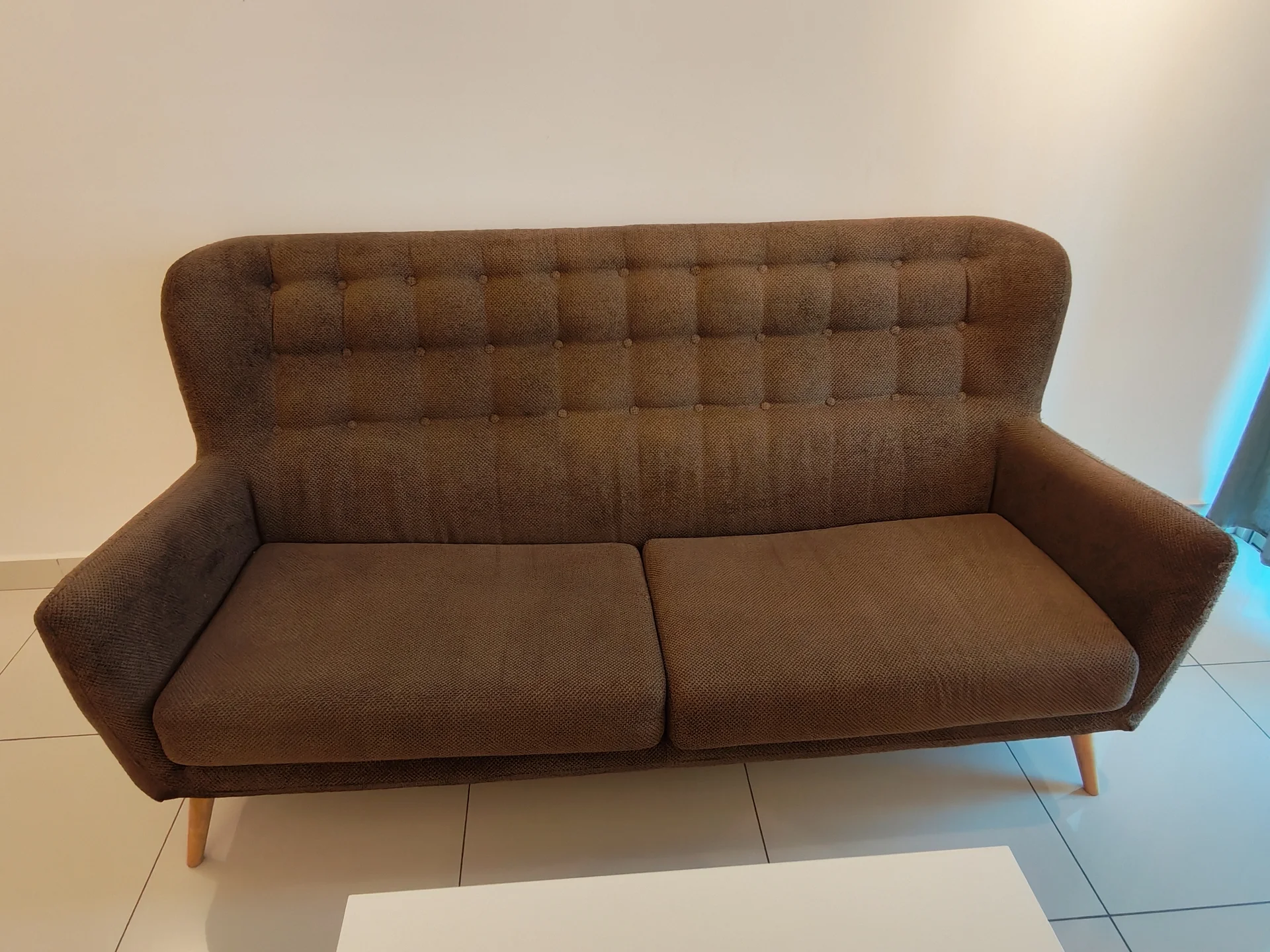 Living features: 3-seater fabric sofa in brown, tufted backrest, wooden legs, white coffee table, white walls, tile flooring, natural light from window, minimalist decor; style: modern.