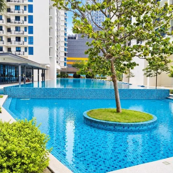 Exterior type: swimming pool deck; features: infinity pool, pool deck with tiled coping, blue mosaic tile pool finish, raised pool edge, circular island planter in pool, tree planted in pool island, surrounding high-rise residential towers, glass-walled clubhouse or pavilion, covered walkway, landscaped shrubs, mature trees, urban skyline background; style: modern.