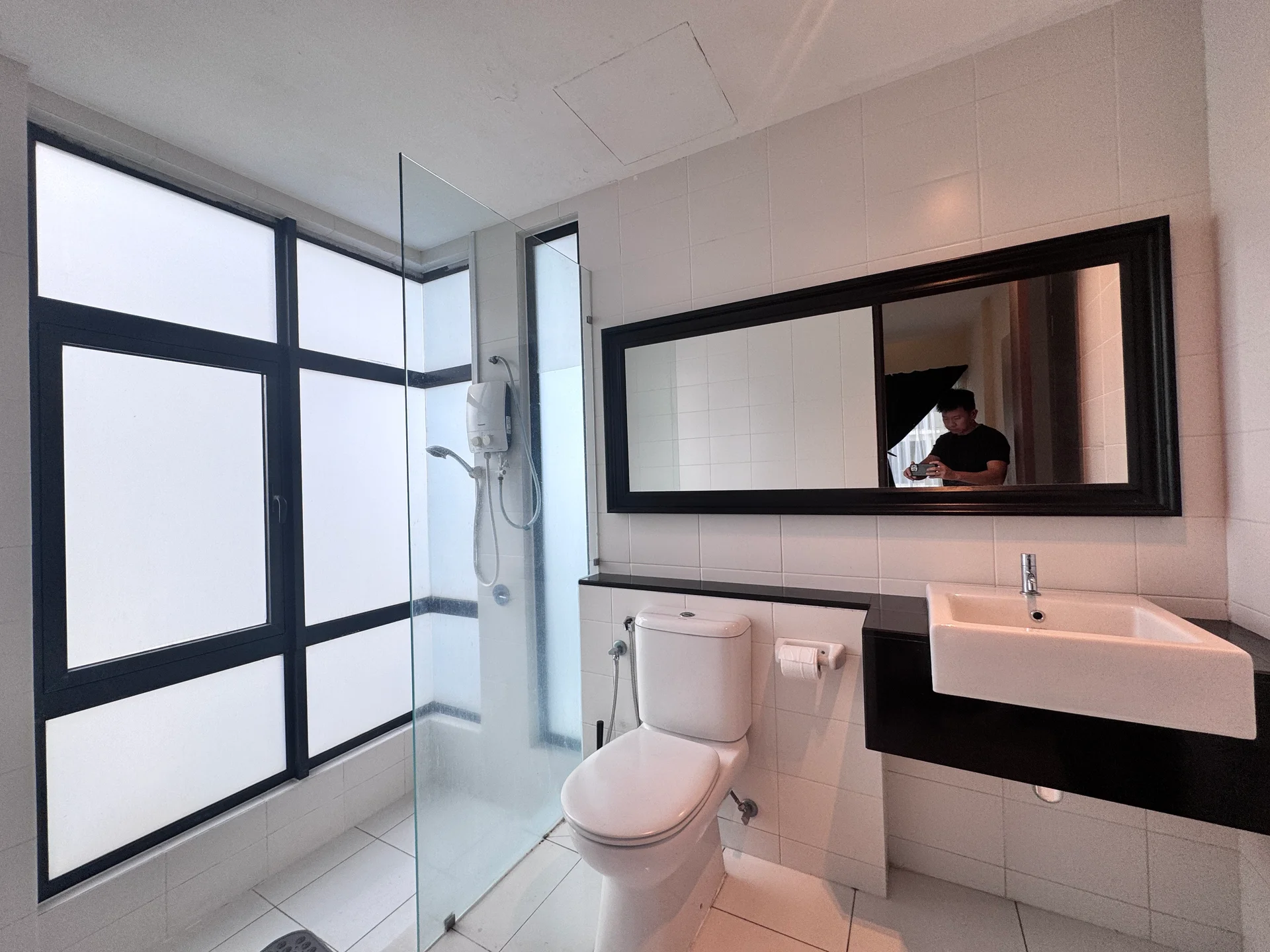 Bathroom features: walk-in shower with glass enclosure, chrome handheld shower, white toilet, single-sink vanity with black countertop, mirror above vanity, white tiled walls, white tiled flooring, floor-to-ceiling windows, black framed windows; style: modern.
