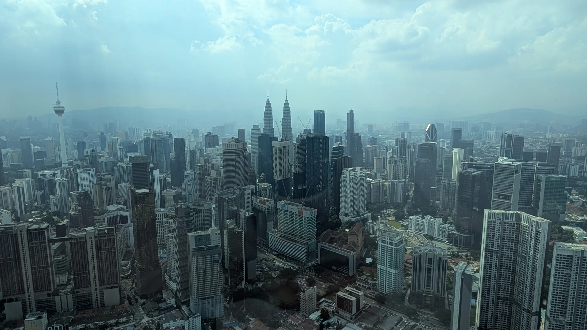 Exterior type: high-rise condominium; features: city skyline view, visible landmarks including Petronas Twin Towers and KL Tower, urban landscape with a mix of skyscrapers and low-rise buildings, cloudy sky, distant mountains, high-rise buildings with glass facades; style: modern.