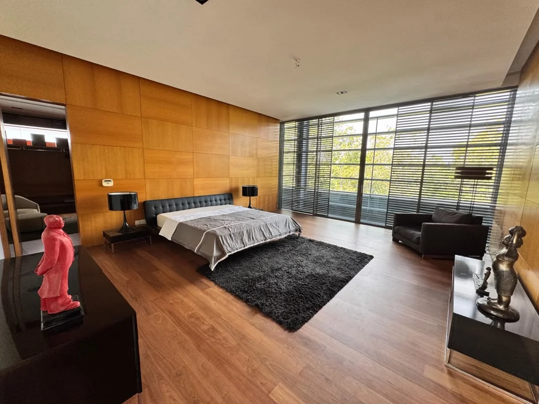 Bedroom features: king-size bed with grey bedding, black leather headboard, 2 bedside lamps with black shades, black fabric sofa, black area rug, wood paneling on walls, large windows with horizontal blinds, wood flooring, decorative statue on side table, decorative figure in red on dresser; style: modern; view: garden.