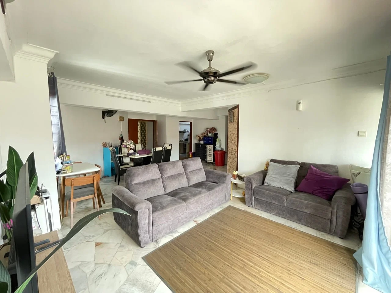 Living features: 3-seater fabric sofa in grey, 2-seater fabric sofa in purple, ceiling fan with light, marble tile flooring, area rug, wall-mounted TV, decorative plants, large windows with sheer curtains, dining table with chairs visible in background, open layout to dining area; style: modern.