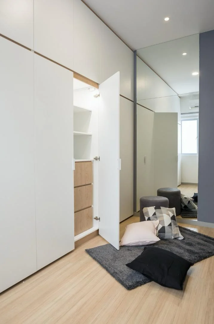 Room features: white cabinetry with sliding doors, wooden shelving inside cabinet, mirrored wall, light wood flooring, recessed ceiling lights, grey area rug, black cushions, decorative pillows with geometric pattern, small ottoman, window with natural light; style: modern.