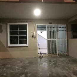 Room features: grey concrete flooring, cream painted walls, large window with white frame, metal security grille door, single light fixture on ceiling.